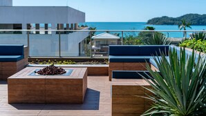 A rooftop terrace with wooden seating, a fire pit, and a view of the ocean and distant hills. - Apto vista mar 4 dormitórios e piscina (Bombinhas)