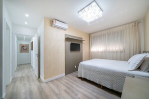 A bedroom with a bed, a flat-screen TV, an air conditioning unit, and a hallway leading to another room.