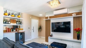 A modern living room with a sofa, ottoman, and a TV mounted on the wall. There are shelves with wine bottles and a small refrigerator. - Apto vista mar 4 dormitórios e piscina (Bombinhas)