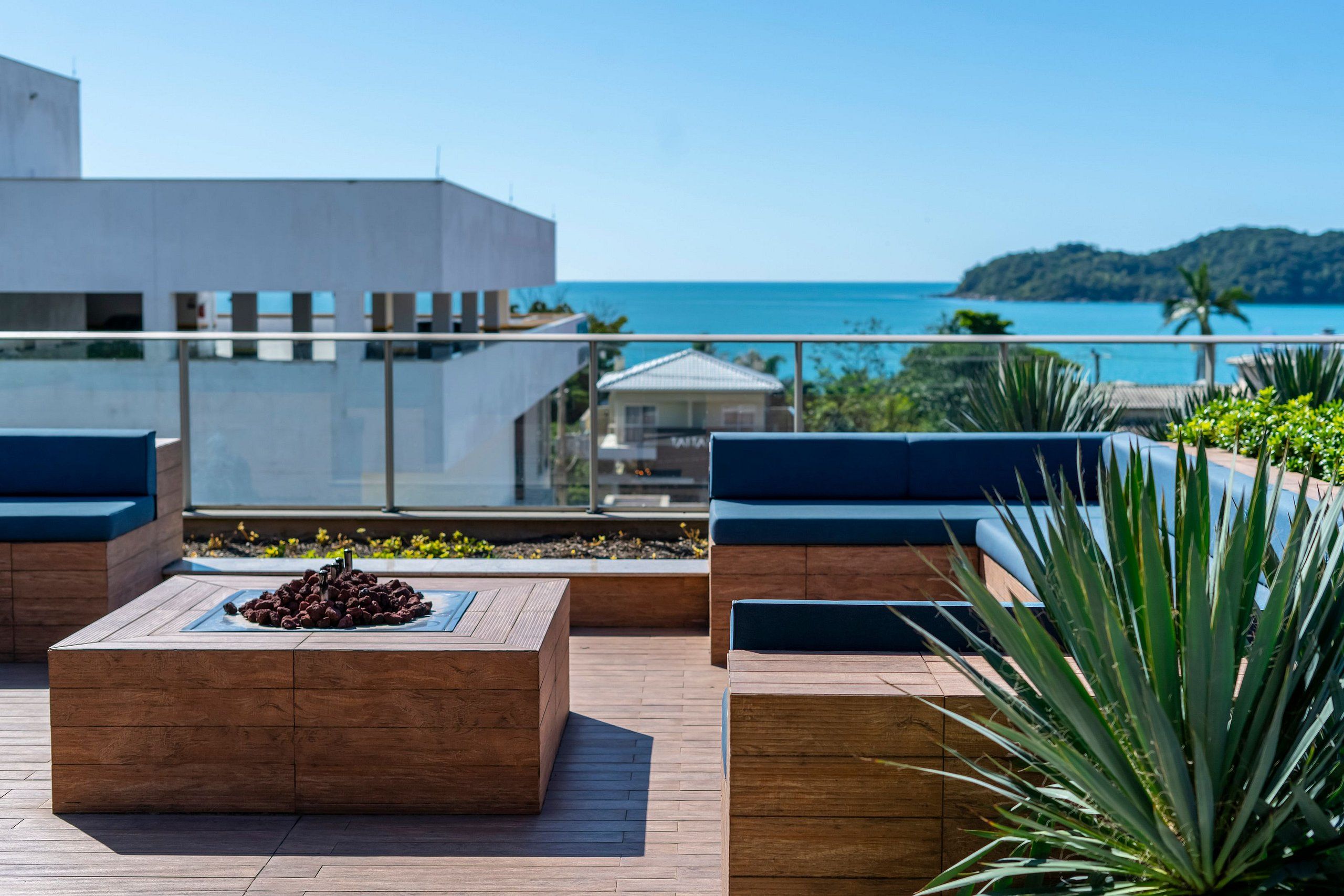 A rooftop terrace with wooden seating, a fire pit, and a view of the ocean and distant hills.