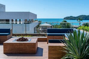 Eine Dachterrasse mit Holzmöbeln, einem Feuerkorb und Blick auf das Meer und die entfernten Hügel.