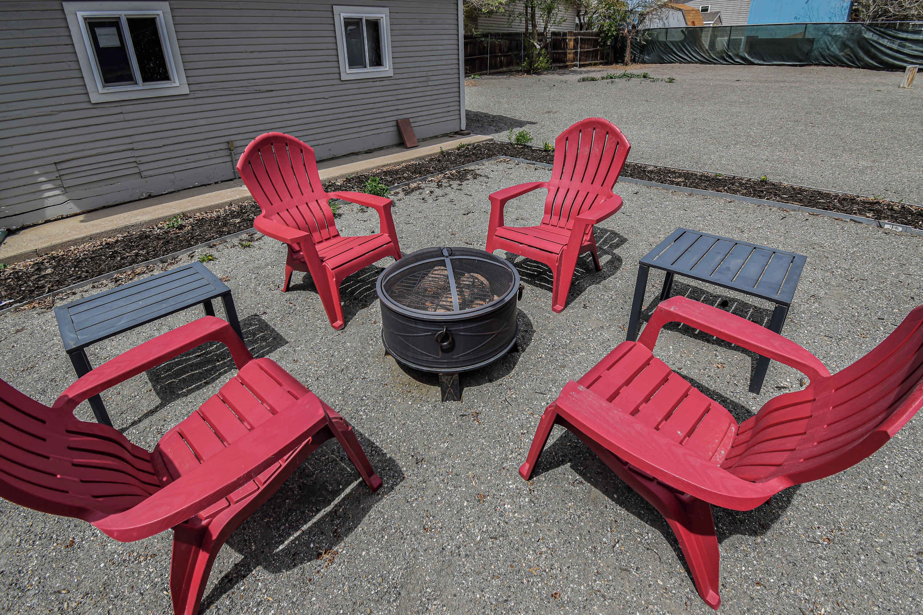 A fire pit surrounded by four red Adirondack chairs and a small table, set on a gravel surface in front of a house.