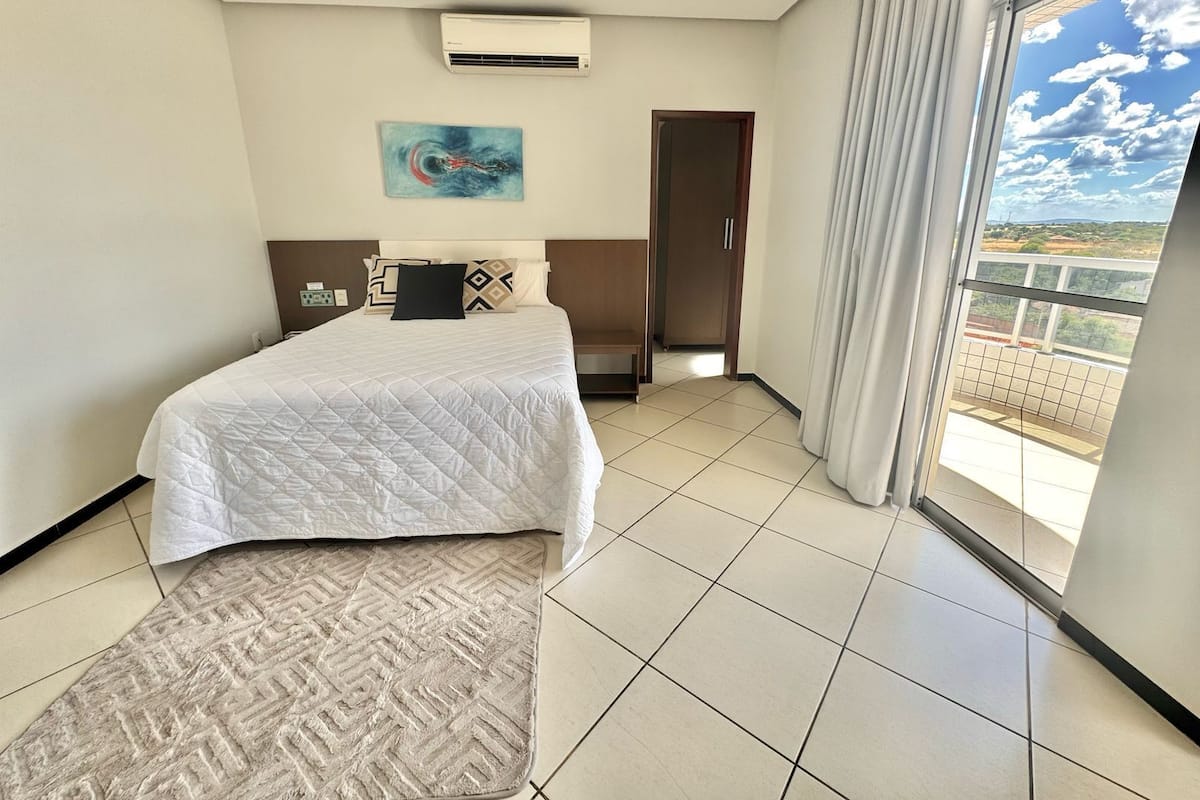 Habitación de hotel con cama, un cuadro, equipo de aire acondicionado, vista por una ventana y puerta que da a otra habitación.