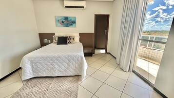A hotel room with a bed, a painting, an air conditioning unit, a view through a window, and a door leading to another room.