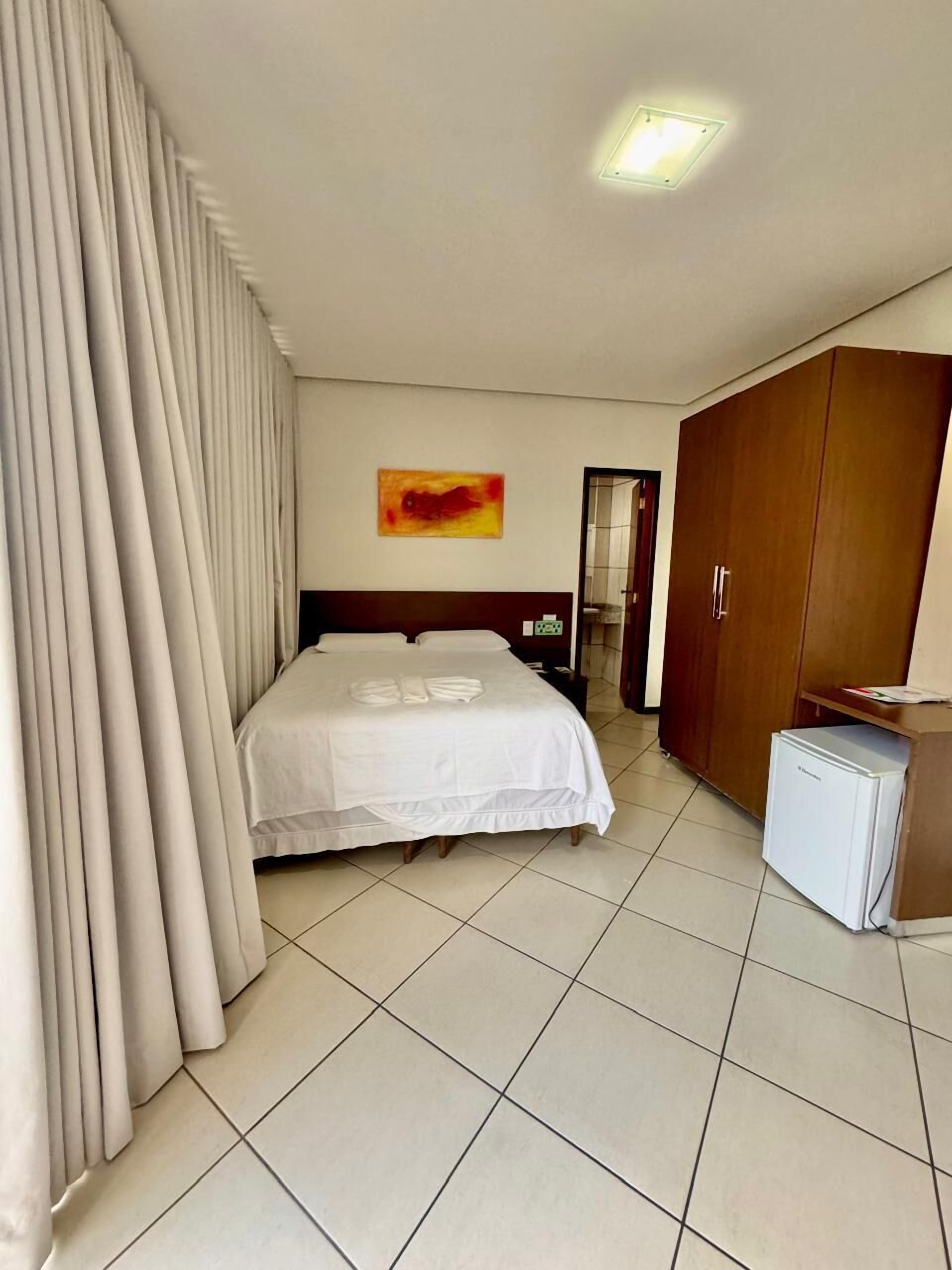 Una habitación de hotel con cama, cortinas, un armario de madera, un refrigerador pequeño y un cuadro en la pared.