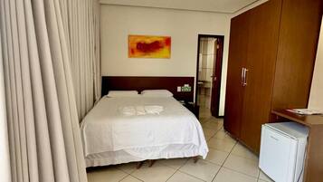 Una habitación de hotel con cama, cortinas, un armario de madera, un refrigerador pequeño y un cuadro en la pared.