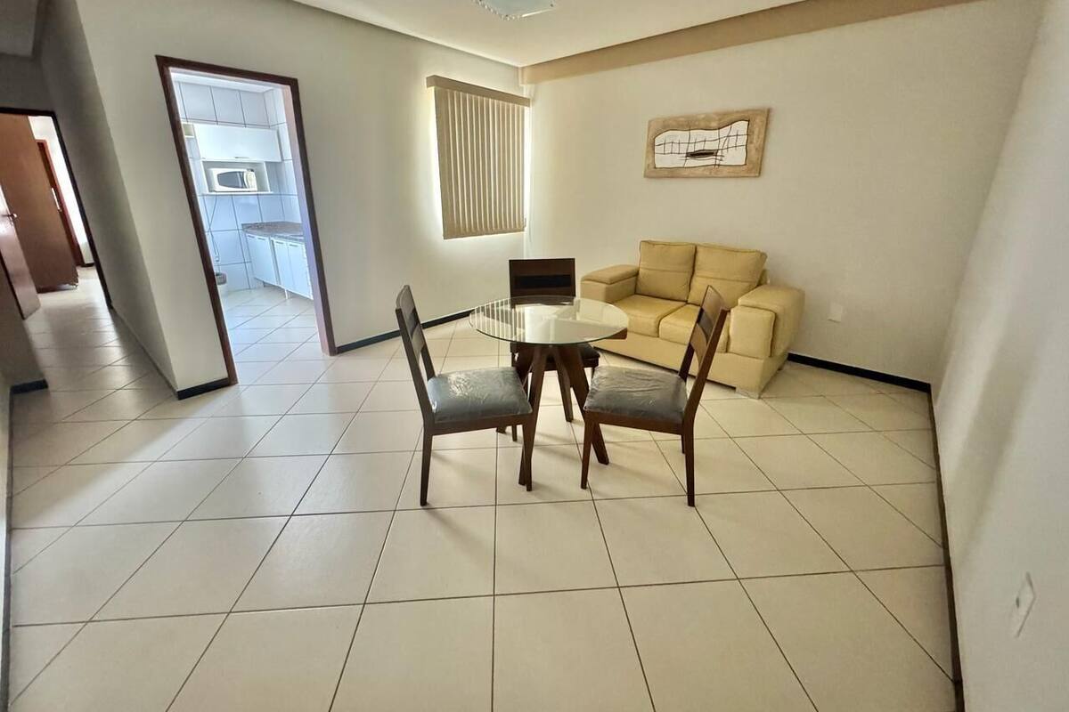 Un salón amplio con una mesa redonda de vidrio y cuatro sillas, un sofá color beige y un ventanal grande con persianas.
