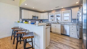 Una cocina moderna con gabinetes blancos, una isla central con taburetes, electrodomésticos de acero inoxidable y vistas a través de las ventanas.