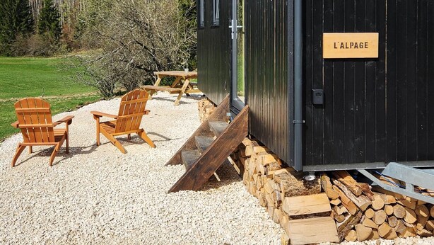 A rustic cabin with a gravel driveway, wooden chairs, and a stack of firewood. - Tiny House L'Alpage (Les Bouchoux)