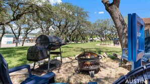 A barbecue setup with a grill and a fire pit in a park-like setting with trees and a blue sign. - LakeTravis Cozy Bungalow 6 (Volente)