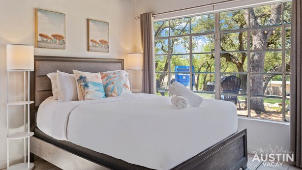 A bedroom with a large bed, two wall-mounted lamps, and a window with a view of trees and a blue sign. - LakeTravis Cozy Bungalow 6 (Volente)