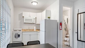 Een kleine keuken met witte kasten, een magnetron, een fornuis en een koelkast. Er is een eettafel met stoelen en een brandblusser aan de muur bevestigd.