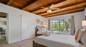 A bedroom with a wooden ceiling, a ceiling fan, two beds, a window with a view of trees, and a door leading to another room. - LakeTravis Bungalow7 (Volente)