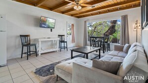 A spacious living room with a wooden ceiling, a large window with a view of trees, a white sofa, a coffee table, and a TV mounted on the wall.