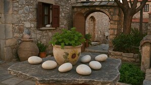 Une table en pierre, agrémentée d’une plante en pot et de quelques pierres, est placée dans une cour bordée de murs en pierre et surmontée d’une arche.