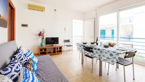 Un soggiorno con divano, TV e tavolo da pranzo con sedie. Include un balcone con ringhiera e vista sui palazzi.