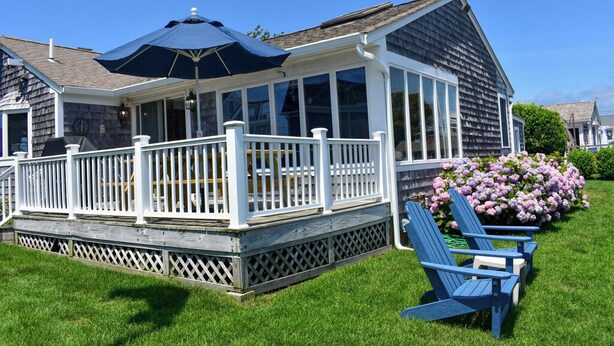 A covered deck with a white railing, a blue umbrella, and blue Adirondack chairs.