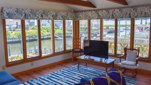 A living room with a TV, a sofa, and a chair, large windows with a view of a water body and buildings.