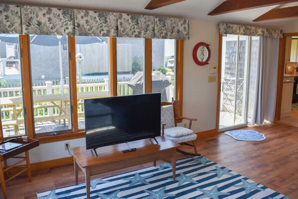 A living room with a TV on a wooden stand, a patterned rug, and a view of a backyard through large windows.