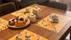 Olohuoneessa on sohva, ruokapöytä, jonka päällä on teekannu, kuppeja ja leivonnaisia. Seinällä on taideteos ja sohvan yläpuolella on taulu.