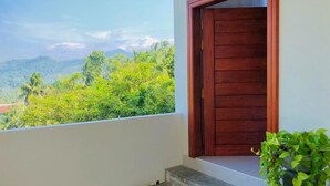 A balcony with a wooden door, a concrete floor, and a view of lush greenery and mountains. - Meridian Holiday Resort (Ratnapura)