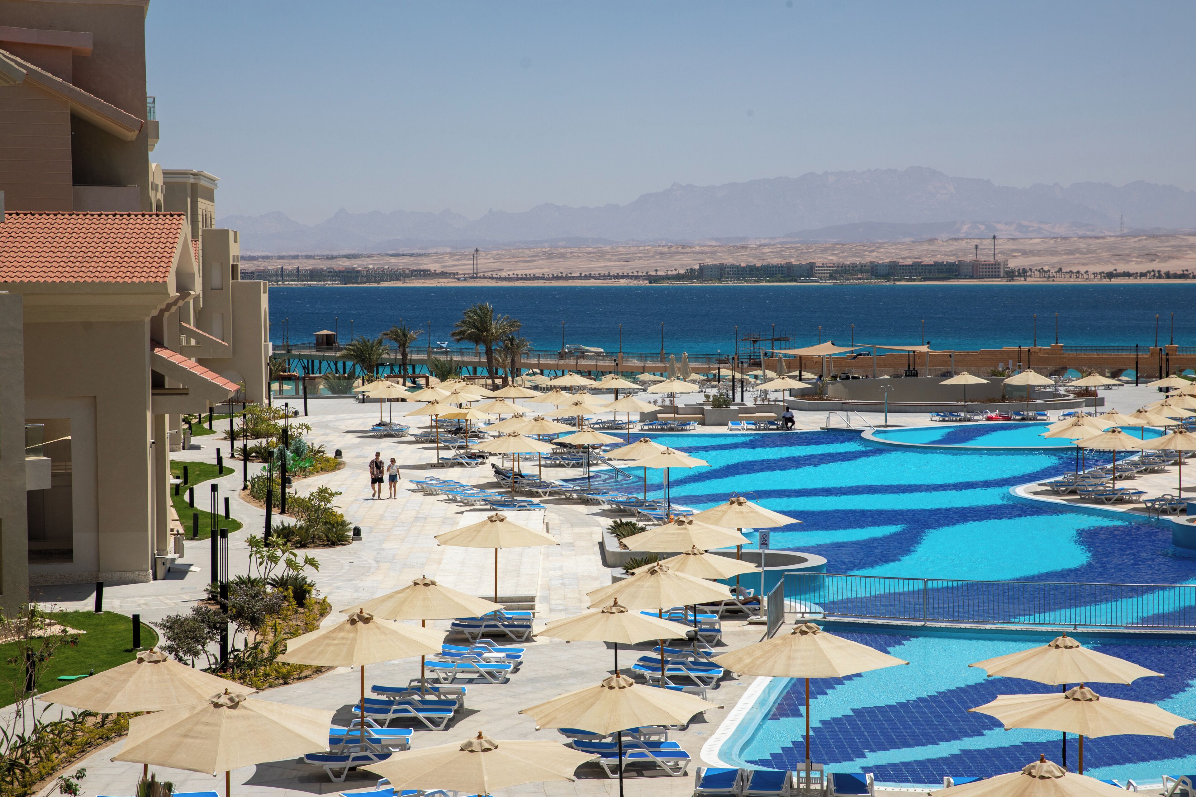 A resort with a swimming pool, sun loungers, umbrellas, and a view of the sea and mountains.