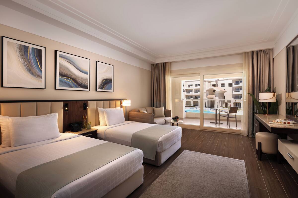 A hotel room with two beds, a desk, a chair, a small table, and a view of a pool and buildings.