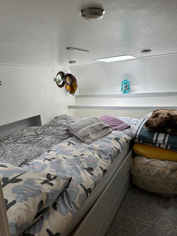 Una habitación compacta y bien iluminada con una cama individual, un edredón estampado, un pequeño estante con un farol y un espejo circular en la pared.