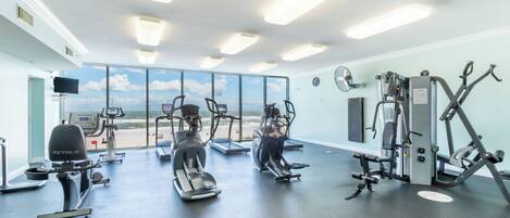 A gym with cardio machines, weight equipment, and large windows offering a view of the beach.