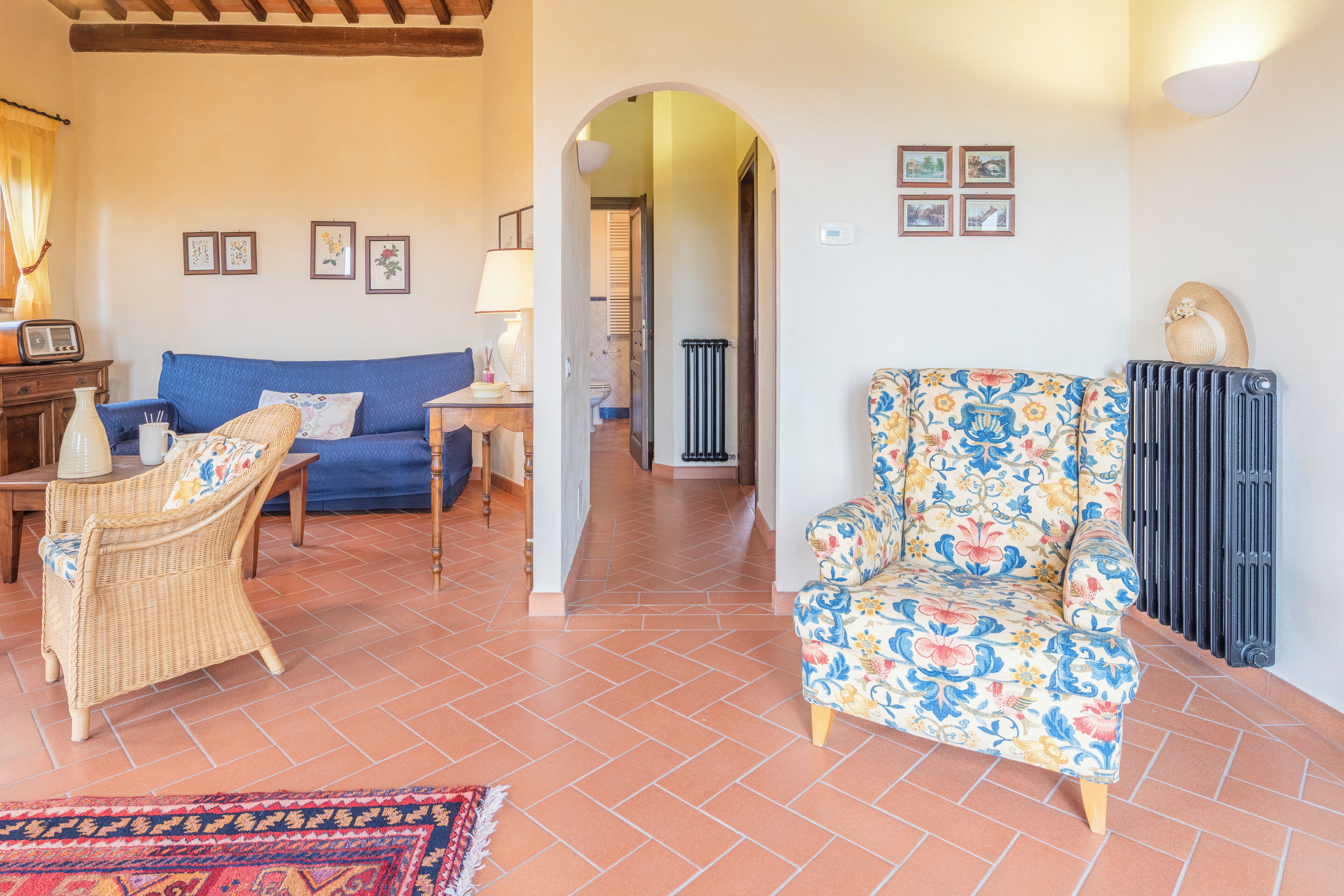 A living room with a blue sofa, wicker armchair, and a floral armchair. There is a tiled floor and a radiator on the wall.