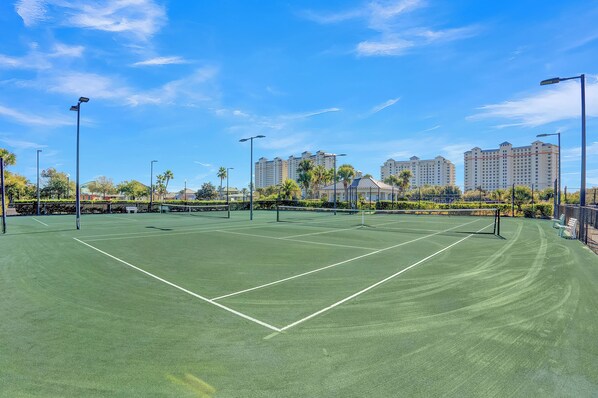 Una cancha de tenis con red, rodeada de palmeras y edificios al fondo.