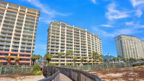 A coastal hotel with multiple floors, a wooden boardwalk leading to the beach, and palm trees.