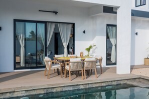 A modern outdoor patio with a pool, wooden furniture, and large sliding glass doors.