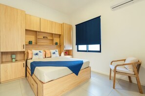 A bedroom with a bed, wooden wardrobe, bedside table, chair, and a window with a blind.