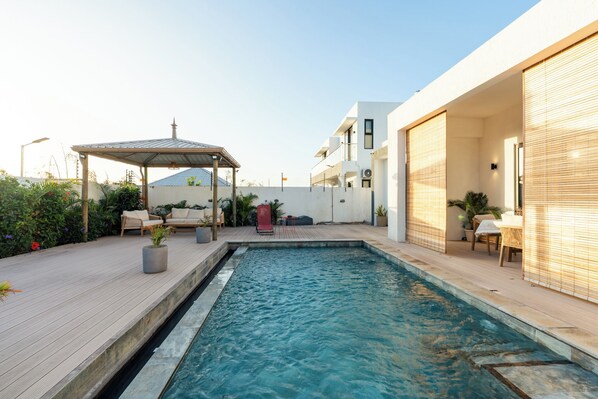 A swimming pool with a wooden deck, a covered seating area, and a modern building with large windows.