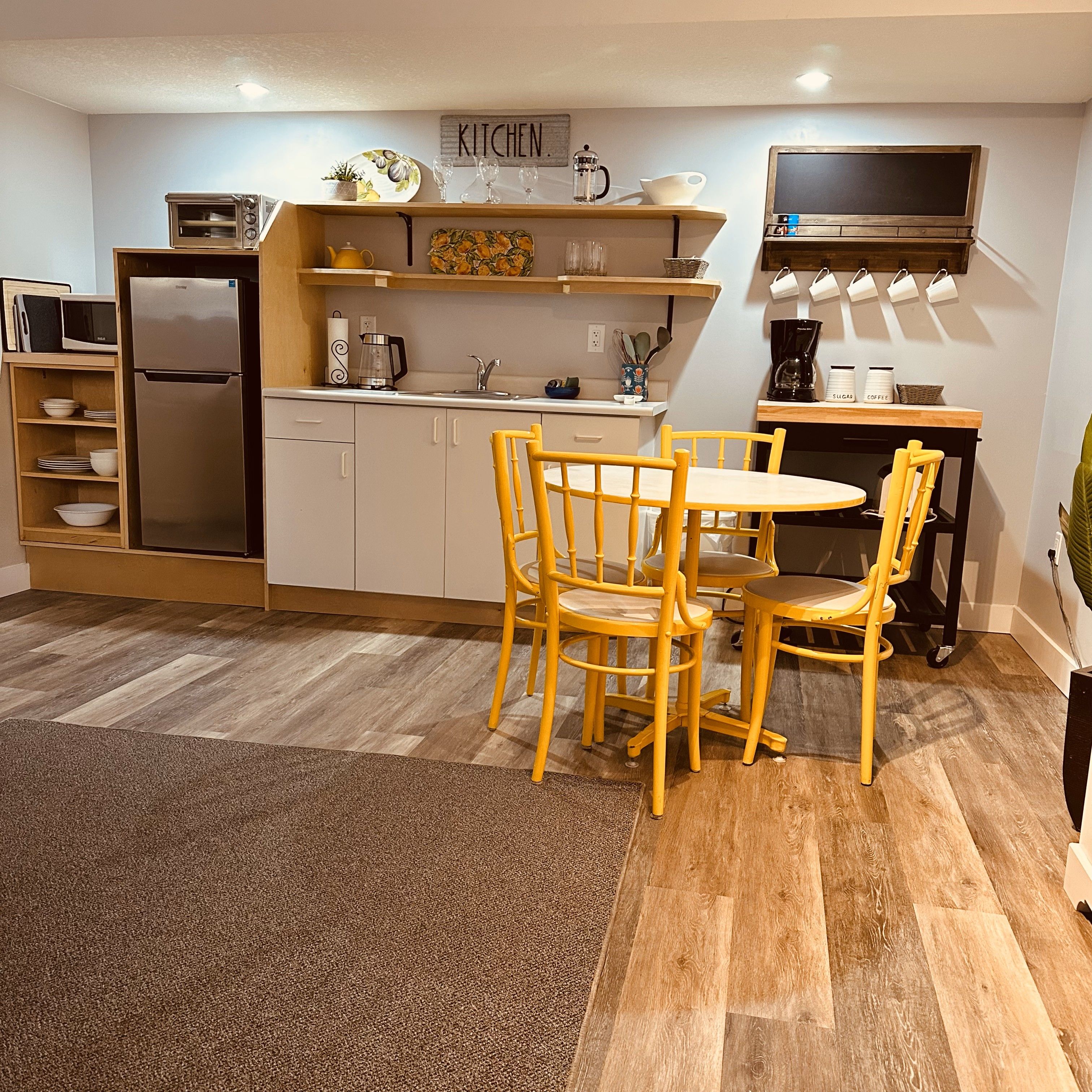 A compact kitchen with a refrigerator, microwave, and coffee maker. There is a dining table with chairs and a rug on the floor.
