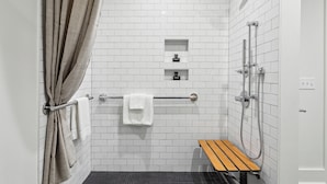 Een moderne badkamer met witte betegelde wanden, een douche met handdouche, een handdoekrek met twee witte handdoeken en een houten bankje.