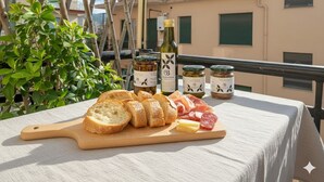 A table with a variety of food items including bread, cheese, and cured meats, accompanied by bottles of olive oil, set on a balcony with a view of buildings and greenery. - Cycling Riviera apartment (Imperia)