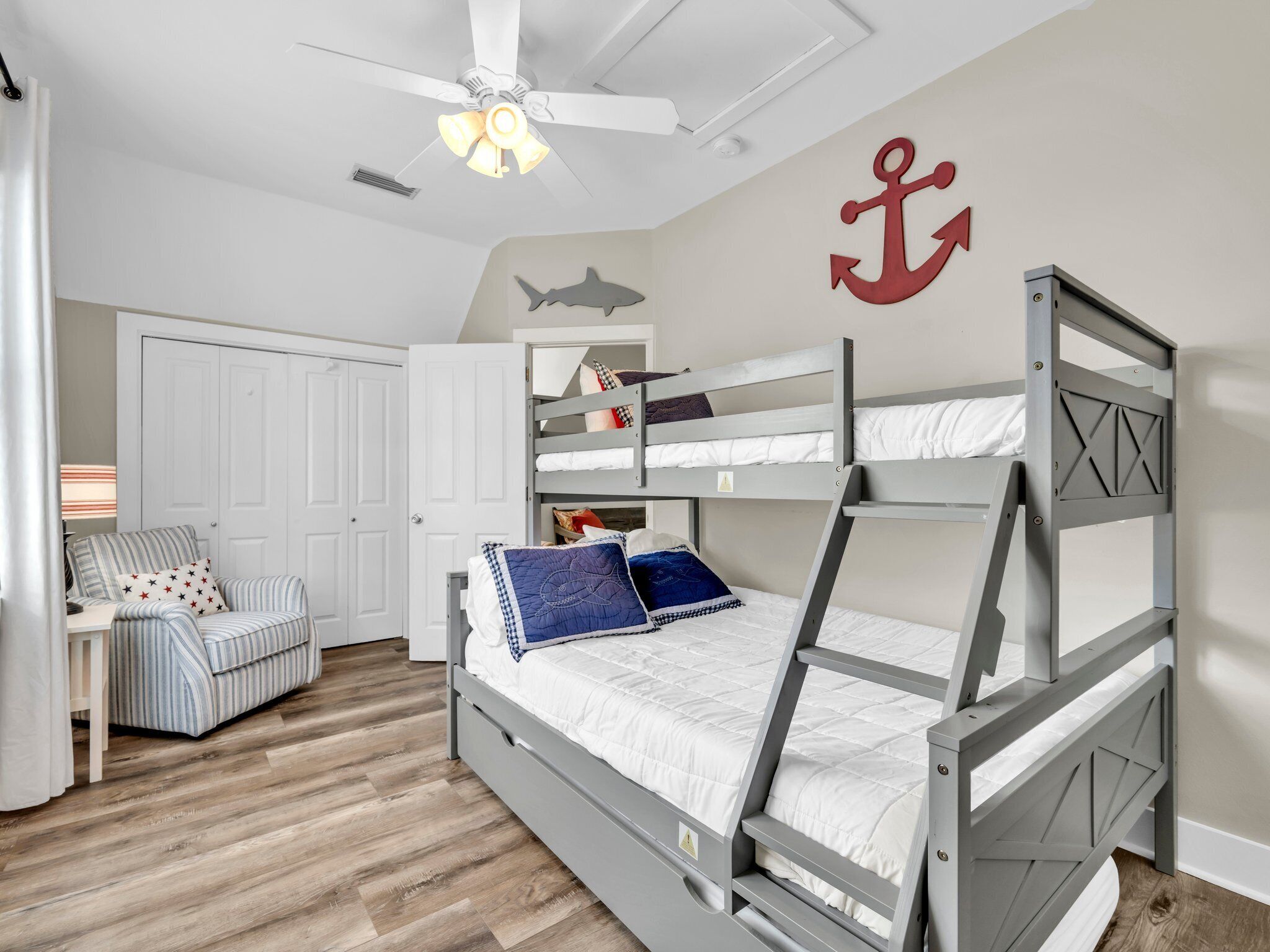 A bunk bed room with a ceiling fan, a striped chair, a desk with a lamp, and a wall with an anchor and shark decoration.
