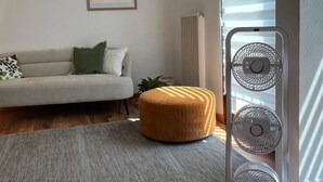 A modern living room with a sofa, a round ottoman, a standing fan, and a framed artwork on the wall.
