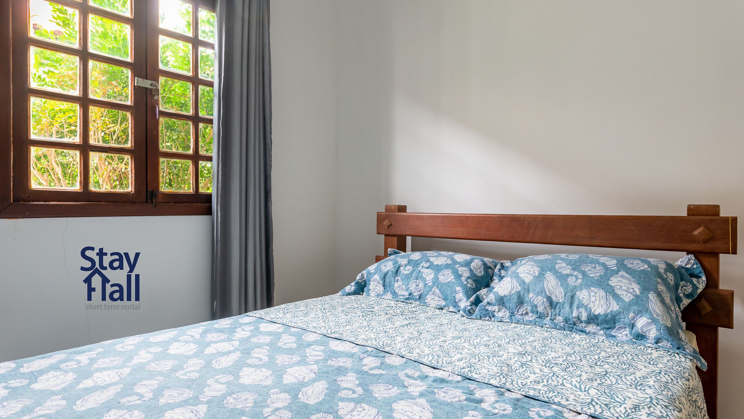 A single bed with a patterned bedspread and pillows, a wooden headboard, a window with a view of greenery, and a 'Stay Hall' sign on the wall.