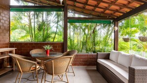 A covered patio with wicker furniture, a brick wall, and large windows overlooking a lush garden. - Casa de Campo Familiar em Gravata 065D (Gravata)