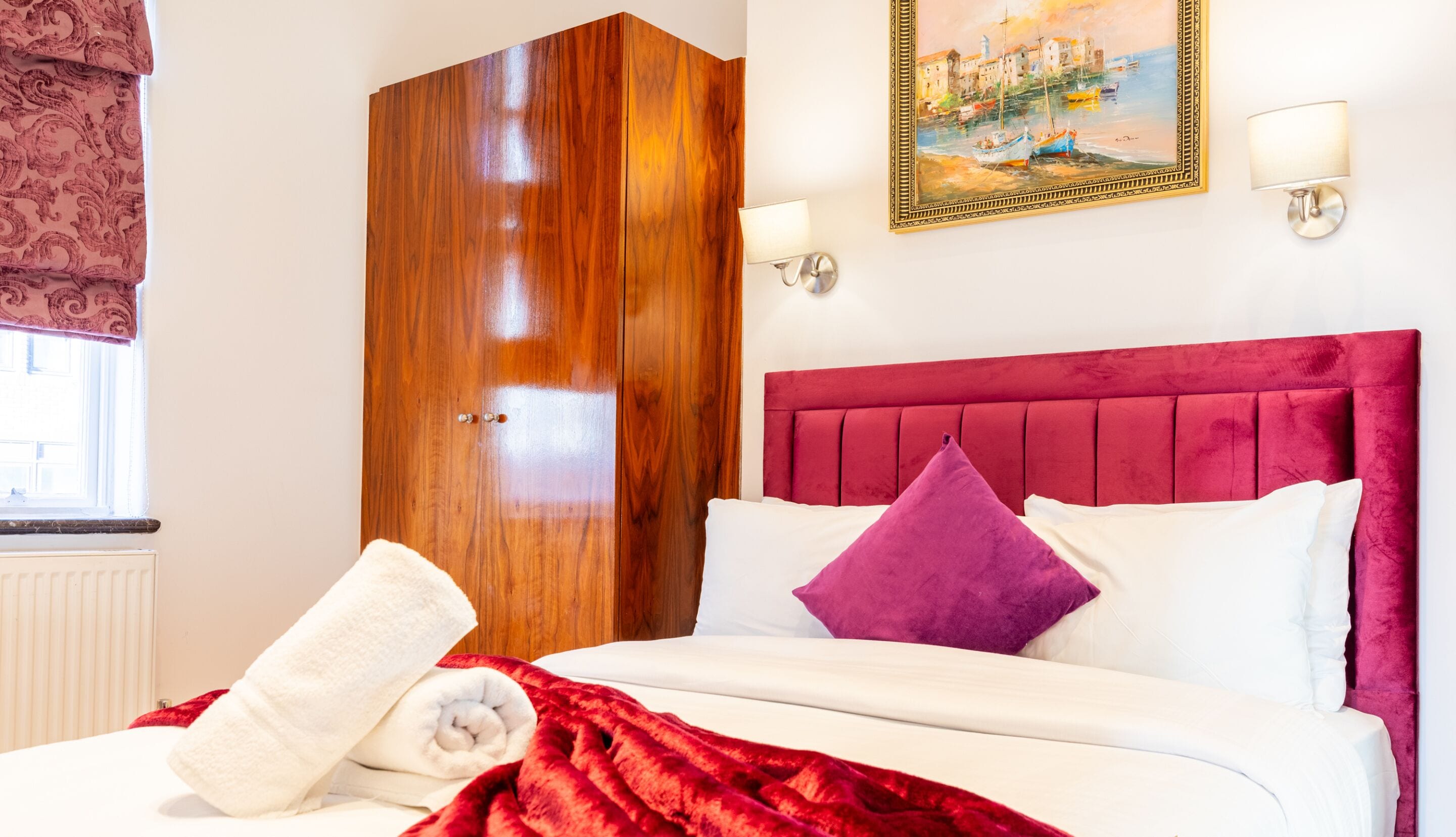 Een hotelkamer met een houten garderobe, een bed met een rode hoofdbord, witte en paarse kussens, een rode deken en een schilderij aan de muur.