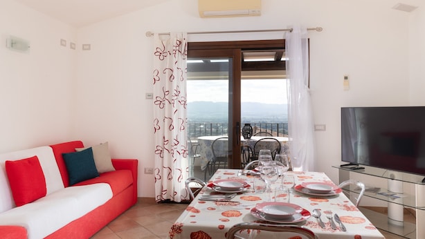 A dining area with a table set for four, a sofa with red cushions, a TV, and a balcony with a view. - Casa Della Vigna 3 by Wonderful Italy (Laerru)