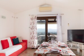 Ein Essbereich mit einem Tisch für vier Personen, einer Couch mit roten Kissen, einem Fernseher und einem Balkon mit Aussicht.