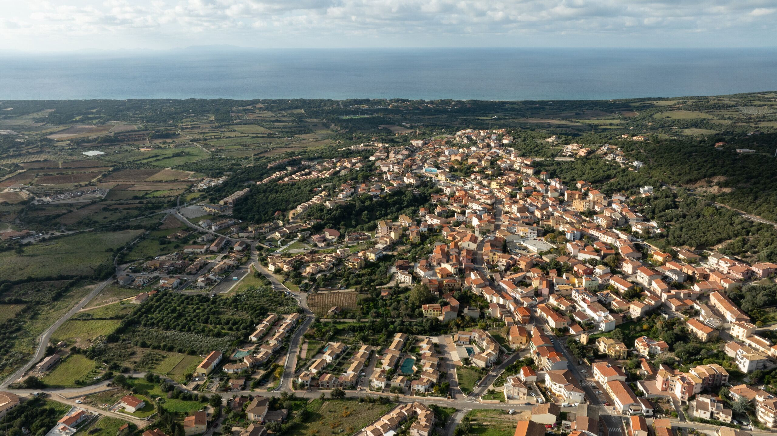 An aerial view of a densely packed residential area with a mix of houses and small buildings, surrounded by green fields and a coastline in the background.