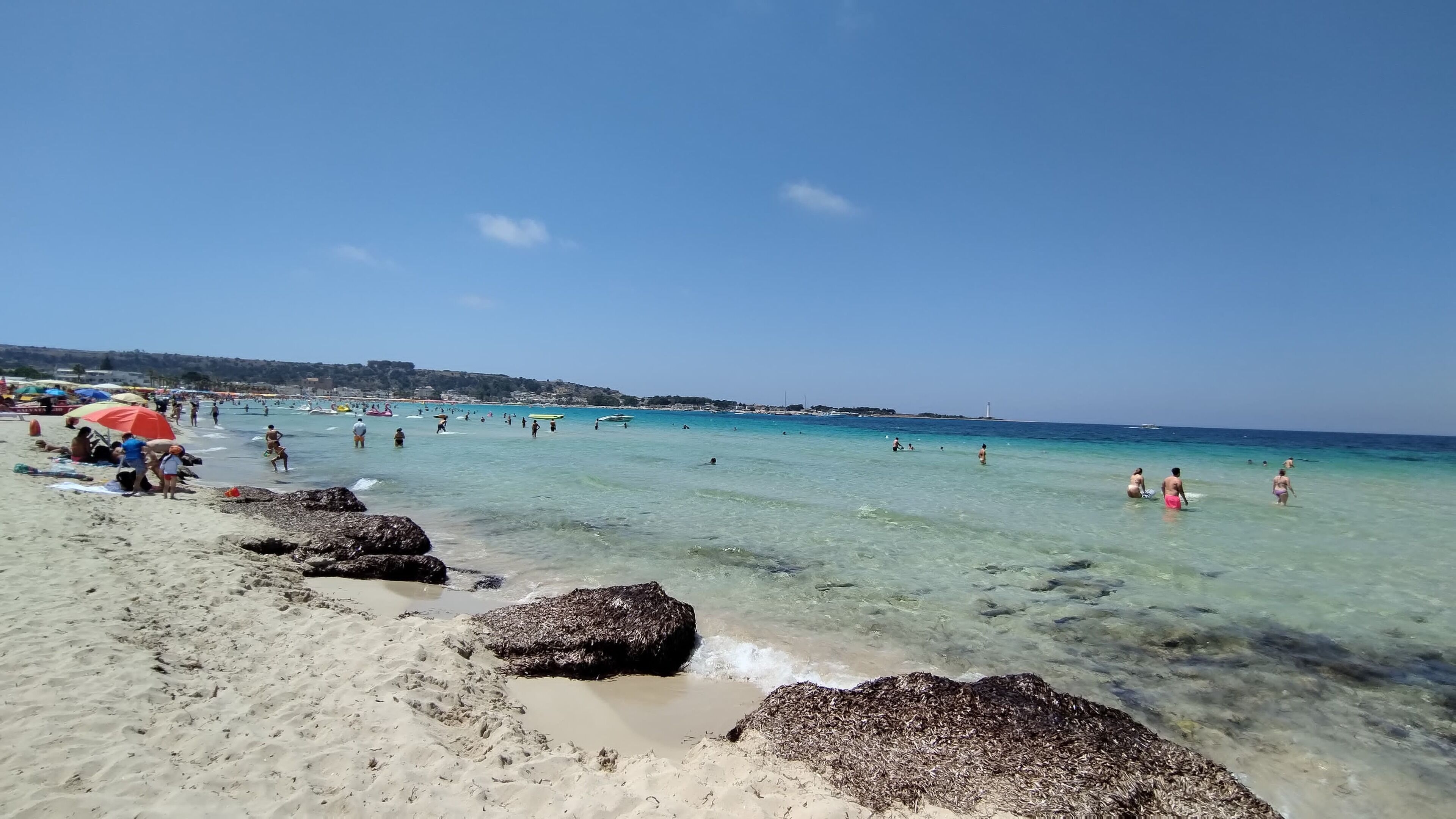 A beach with clear turquoise water, people swimming and sunbathing, and a lighthouse in the distance.