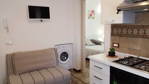 A compact living space with a sofa, washing machine, kitchenette, and a TV mounted on the wall. - DE MEO GUEST HOUSE APPARTAMENT (Castellammare del Golfo)