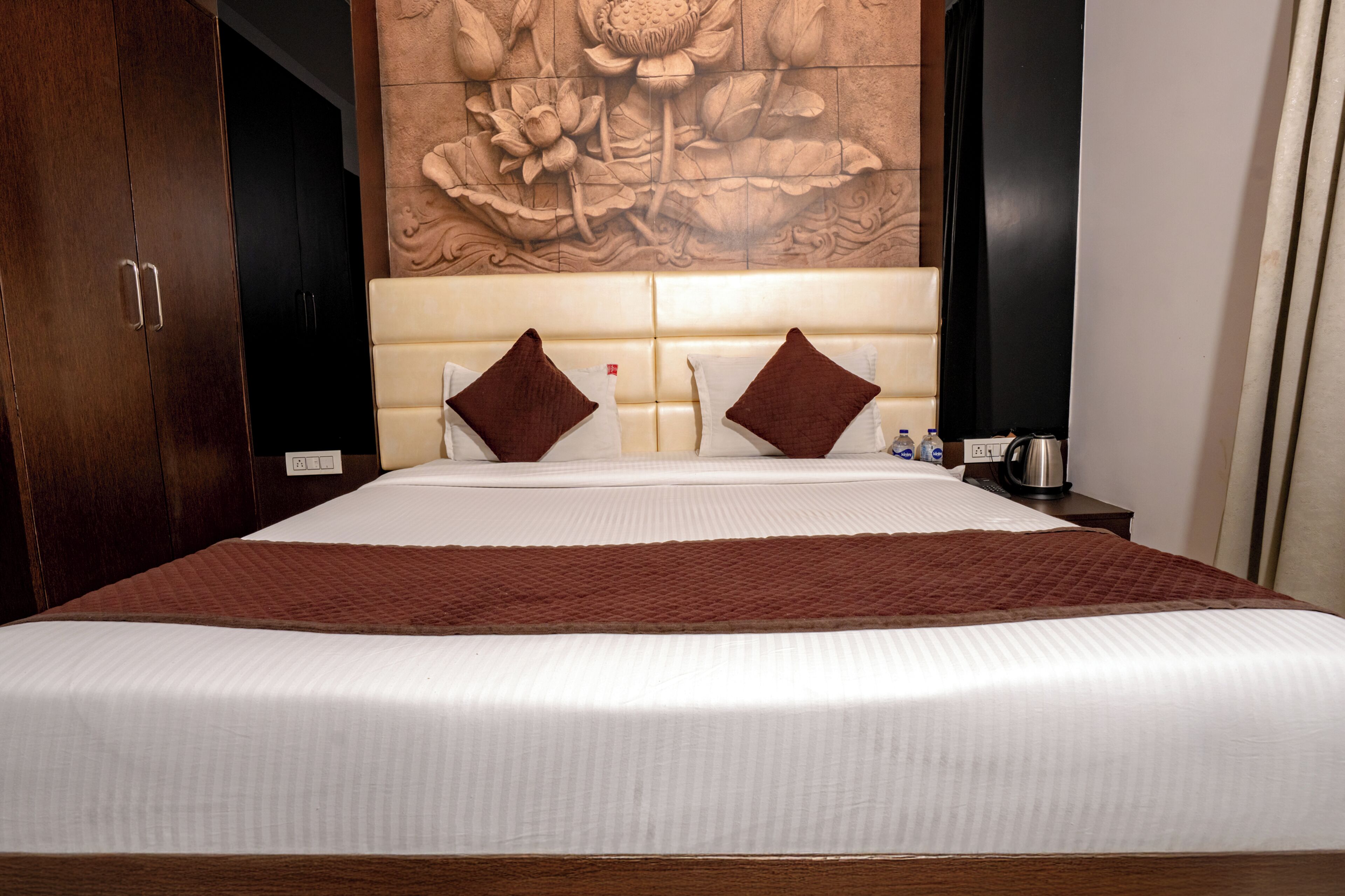 Una habitación de hotel con una cama grande, dos almohadas, un cabecero con diseño floral, un armario de madera y una mesita de noche con hervidor.