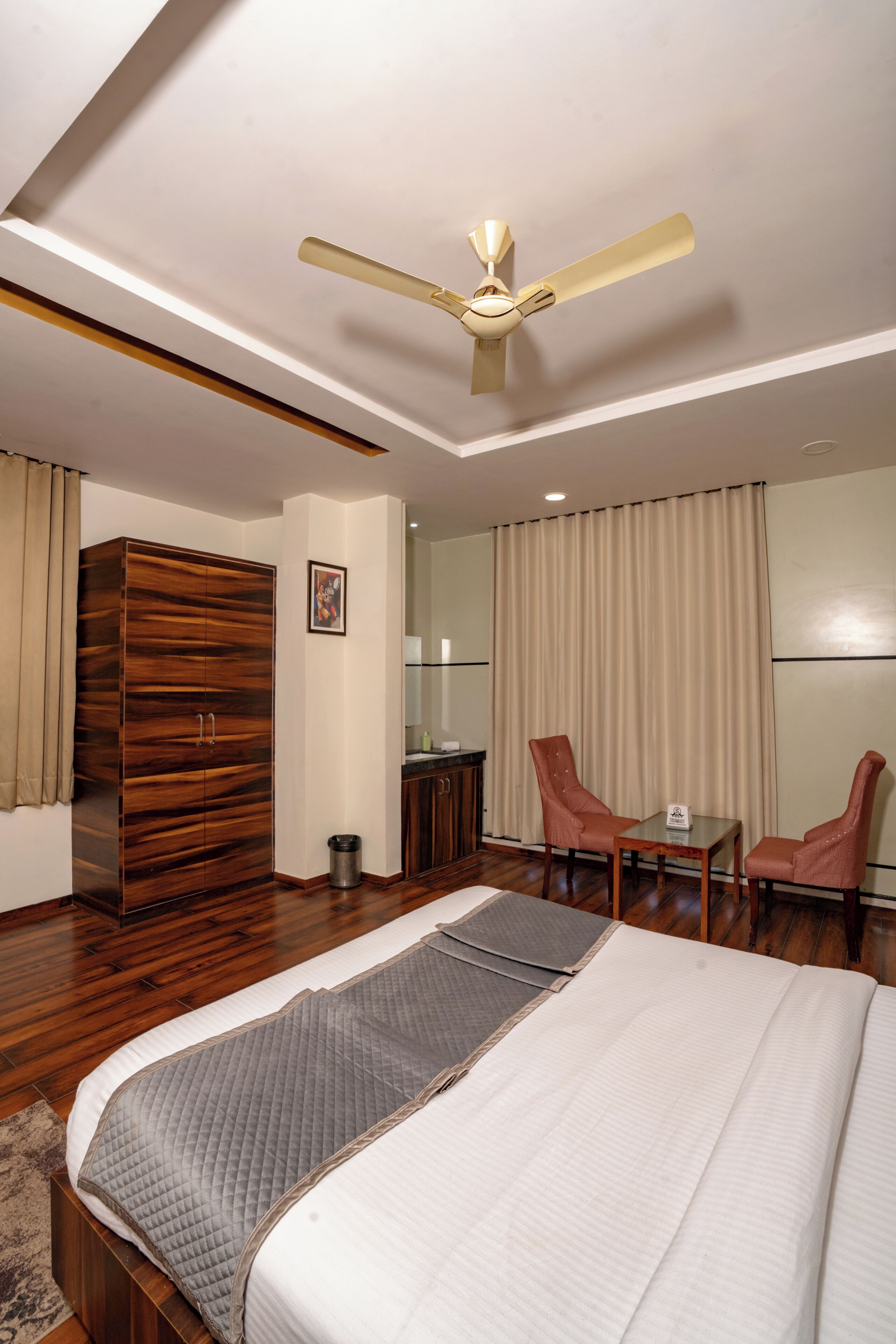 A bedroom with a large bed, a wardrobe, a desk with two chairs, a ceiling fan, and a large window with curtains.